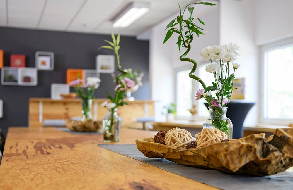 Heller Innenraum mit einem langen Holztisch, Vasen mit Blumen und dekorativen Kugeln als Tischdekoration.