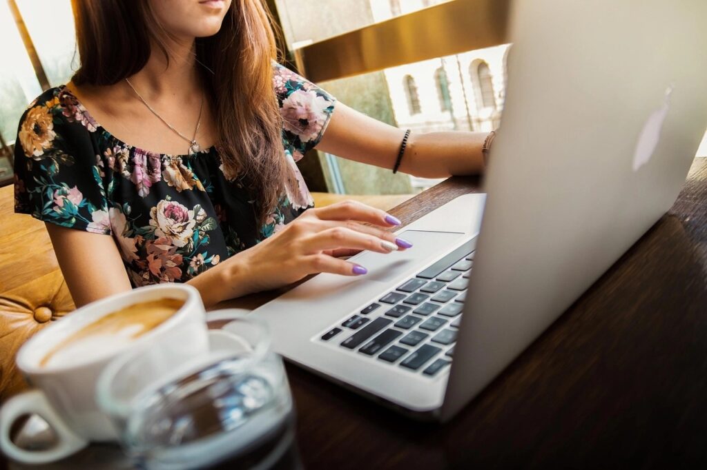 Junge Frau tippt auf einem Laptop an einem Café-Tisch, neben einer Tasse Kaffee.