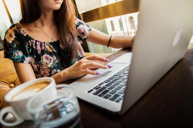 Junge Frau tippt auf einem Laptop an einem Café-Tisch, neben einer Tasse Kaffee.