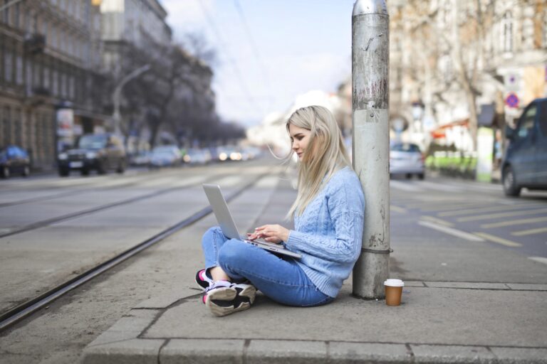 Junge Frau sitzt auf dem Gehweg nahe Straßenbahnschienen und arbeitet an einem Laptop, ein Pappbecher steht daneben.